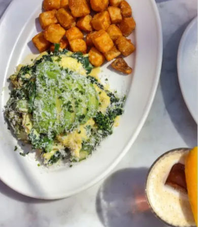 Brunch plate with creamy scrambled eggs topped with sliced avocado and grated cheese over sautéed greens, crispy cubed breakfast potatoes, and a frothy coffee drink with citrus garnish on a marble table.