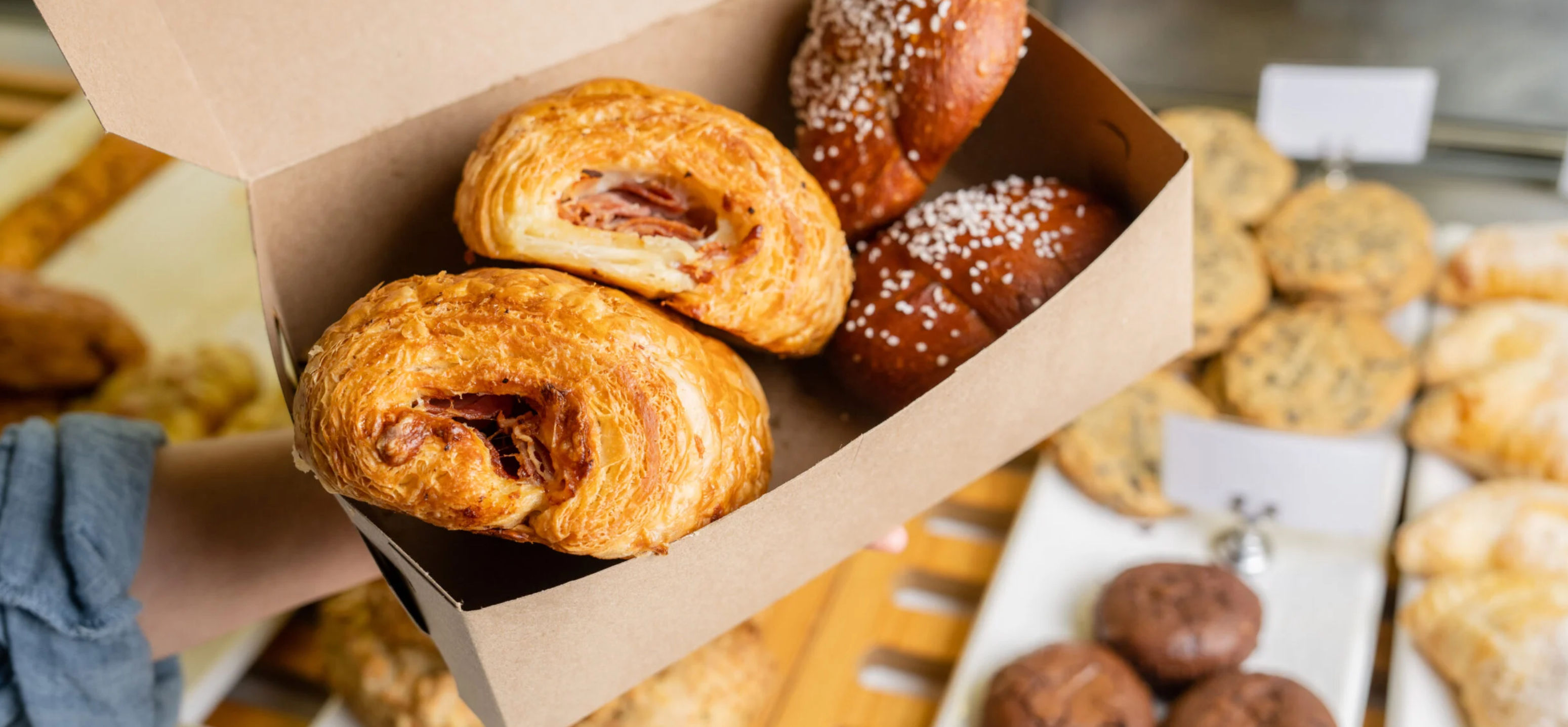 Takeout kraft box holding two flaky savory ham croissants and two sugar-topped brioche rolls over a bakery pastry display with cookies and other pastries