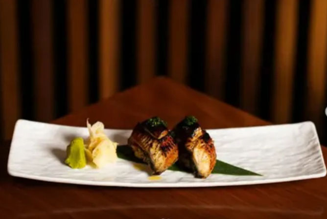 Two pieces of grilled eel (unagi) nigiri on a green leaf with wasabi and pickled ginger, served on a long white rectangular plate on a wooden table in a dimly lit Japanese-style setting.
