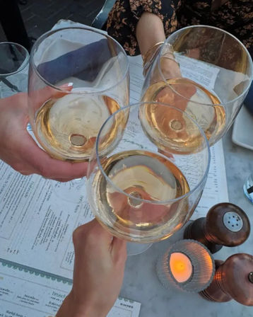 Three hands clinking glasses of rosé wine over menus on a marble outdoor bistro table with candlelight and wooden pepper mills — casual patio cheers.