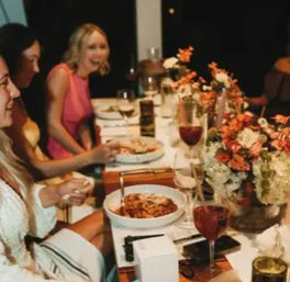 Group of friends laughing at a cozy indoor dinner party around a long table set with pasta bowls, red wine glasses, and colorful floral centerpieces.