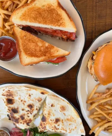 Three enamel plates on a wooden table: a toasted tomato sandwich with fries and ketchup, a cheeseburger with fries, and a charred quesadilla with pickles and greens.