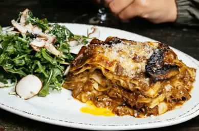 Close-up of oven-browned lasagna layered with meat ragu and melted cheese, served with a peppery arugula and shaved mushroom salad on a white plate — rustic Italian-style dish.