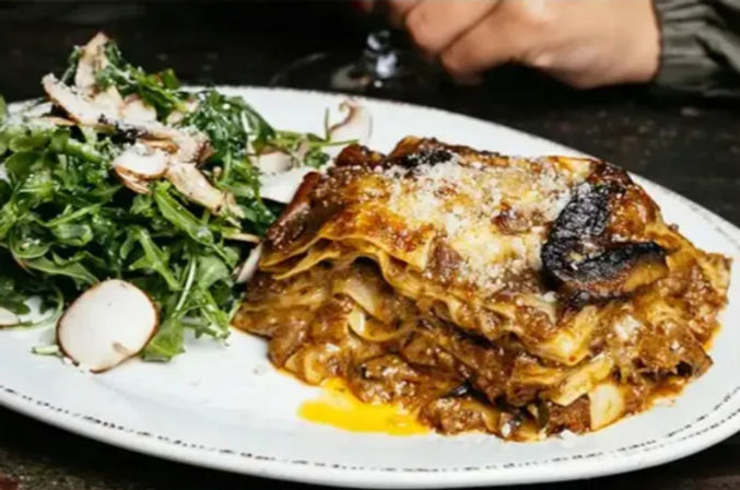 Close-up of oven-browned lasagna layered with meat ragu and melted cheese, served with a peppery arugula and shaved mushroom salad on a white plate — rustic Italian-style dish.