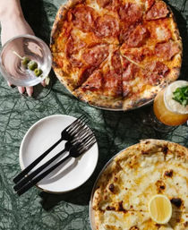 Top-down shot of two pizzas on a green marble table: sliced pepperoni pizza with charred crust and a gooey white cheese pizza with a lemon wedge, accompanied by a martini with olives, a frothy cocktail, and a plate holding three black-handled forks.