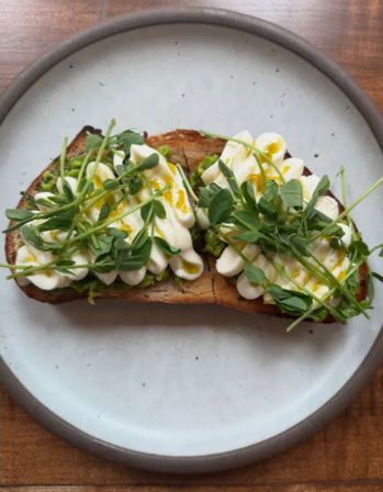 Artisan sourdough toast topped with whipped ricotta, fresh pea shoots and a drizzle of olive oil on a ceramic plate — bright cafe-style brunch