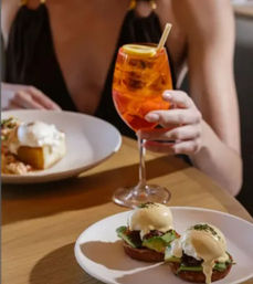 Brunch at a cafe: two avocado eggs Benedict on toasted muffins on a white plate, with a bright orange aperitif garnished with lemon held by a diner.