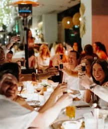 Group dining at a candlelit restaurant — smiling friends around a long table raising small glasses in a lively evening toast.