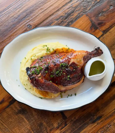 Roasted half chicken with creamy mashed potatoes, chive garnish and green herb sauce on a white plate over a rustic wooden table.