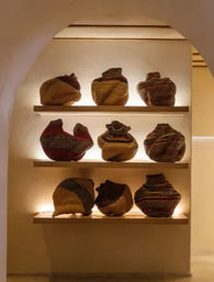 Arched wall niche with warm backlighting showcasing nine handcrafted woven baskets on three wooden shelves — textured, colorful sculptural baskets in a cozy gallery-style display