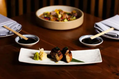 Two pieces of grilled unagi (eel) nigiri with wasabi and pickled ginger on a rectangular plate, with soy sauce dishes, chopsticks and napkins on a wooden table in a Japanese restaurant setting.