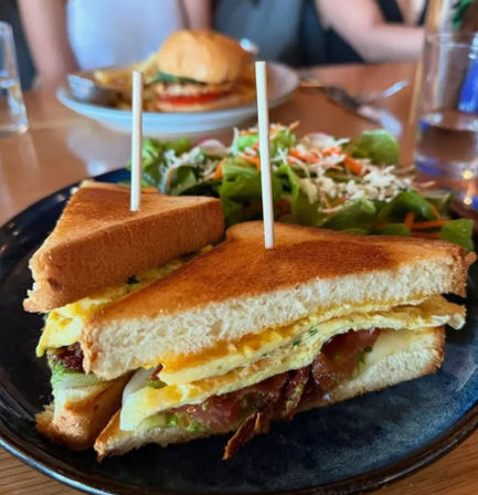 Toasted breakfast sandwich with folded egg, melted cheddar, bacon, lettuce and tomato on white bread, skewered and served with a mixed green salad on a dark plate — brunch-ready café meal.