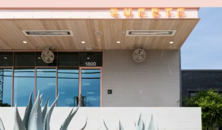 Pink-trimmed modern storefront with glass windows, overhead fans and neon letters, set behind a white wall with sculptural agave plants — desert-inspired exterior.