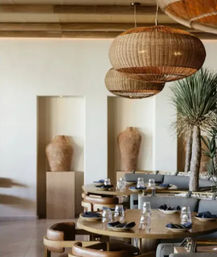 Sunlit Mediterranean-style dining area with round wooden tables set with glassware and navy napkins, curved leather chairs, oversized woven rattan pendant lights, tall terracotta urns in wall niches and a potted palm — relaxed boho-chic interior.
