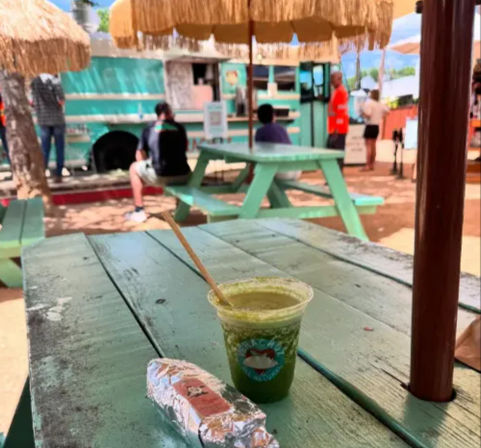 Close-up of a green smoothie in a clear cup with a wooden straw and a foil-wrapped wrap on a weathered turquoise picnic table at an outdoor food-truck seating area with thatched umbrellas.