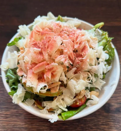 Top-down shot of a fresh mixed-green salad in a white bowl on a wooden table, piled with shaved hard cheese, a dusting of pink seasoning and visible cherry tomatoes — bright healthy lunch bowl.