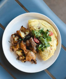 Brunch plate: fluffy omelette topped with cilantro and cherry tomato halves, served with crispy roasted potatoes on a white plate over a blue table