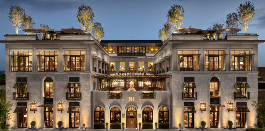 Illuminated luxury boutique hotel facade at dusk with symmetrical arched entrances, tall windows, rooftop terraces with trees, and warm glowing exterior lighting.