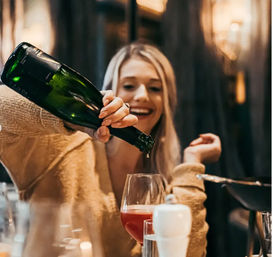 Smiling woman in a cozy sweater pouring red wine from a green bottle into a glass at a warm, candlelit restaurant table.