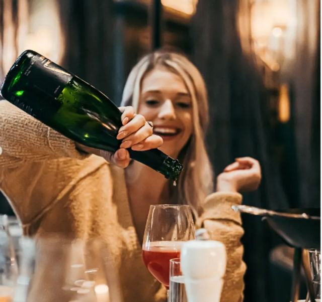 Smiling woman in a cozy sweater pouring red wine from a green bottle into a glass at a warm, candlelit restaurant table.