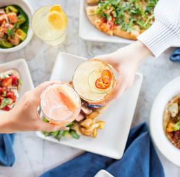 Hands clinking citrus cocktails — one garnished with lime and sliced jalapeño — over a marble brunch table with pizza, salads, and small plates, casual dining cheers