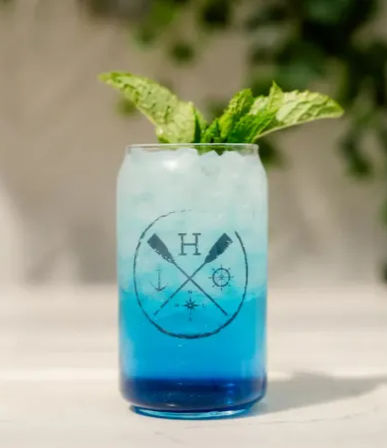 Bright layered blue cocktail in a tall nautical-themed glass with crushed ice and a fresh mint sprig, styled on a light outdoor table