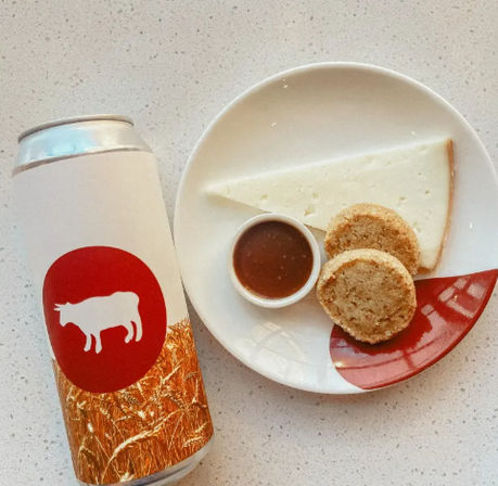 Tall canned drink with cow logo next to a white plate of cheese and crackers — triangular wedge of white cheese, two round crumbly biscuits and a small ramekin of brown chutney on a speckled countertop.