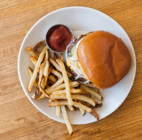 Brioche cheeseburger with pickles and onions, hand-cut fries and a side of ketchup on a white plate atop a wooden table — classic diner-style burger and fries.
