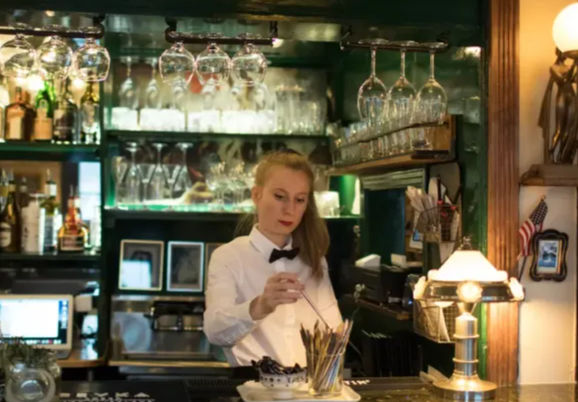 Server in a white shirt and black bow tie stirring a cocktail behind a cozy neighborhood pub bar with hanging wine glasses, back-bar bottles and warm lamp light.