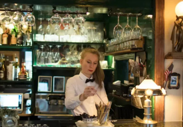 Server in a white shirt and black bow tie stirring a cocktail behind a cozy neighborhood pub bar with hanging wine glasses, back-bar bottles and warm lamp light.