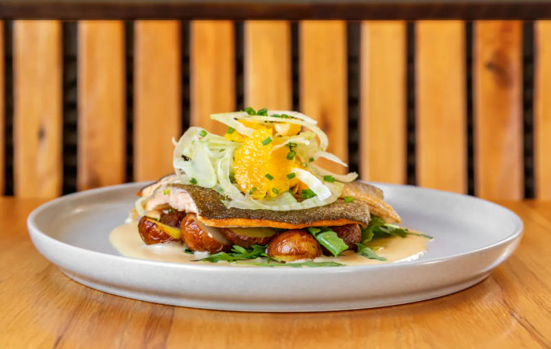 Pan-seared fish fillet with crispy skin on roasted baby potatoes and arugula, drizzled with creamy sauce and topped with shaved fennel, orange segments and chives on a white plate on a wooden table.