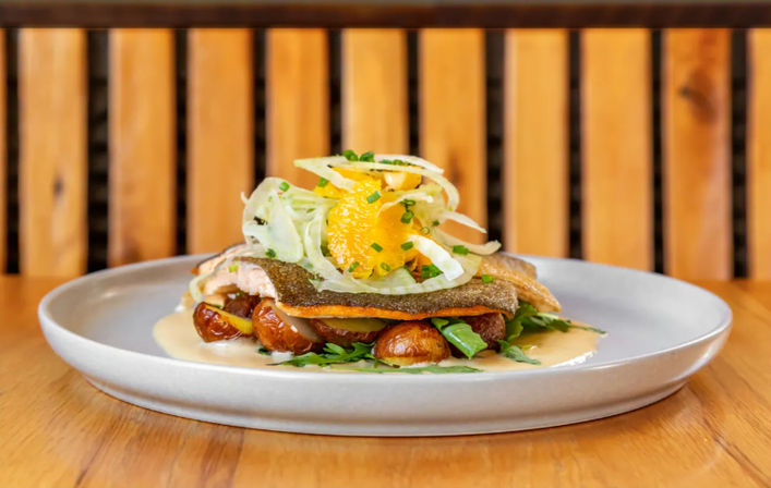 Pan-seared fish fillet with crispy skin on roasted baby potatoes and arugula, drizzled with creamy sauce and topped with shaved fennel, orange segments and chives on a white plate on a wooden table.