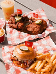 Crispy fried chicken sandwich topped with a pickle, crinkle-cut fries and extra fried chicken on red-and-white checkered paper with an iced drink on an outdoor picnic table
