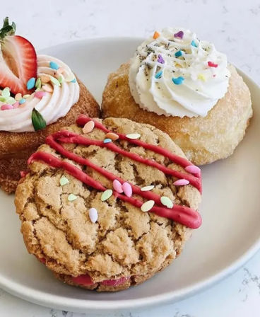 Close-up of a white plate with three colorful bakery-style treats: a cracked oatmeal cookie drizzled with pink icing and pastel sprinkles, a sugar-coated cream-filled donut with whipped frosting and confetti sprinkles, and a frosted donut topped with a halved strawberry.