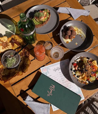 Al fresco brunch on a wooden patio table with avocado toast, a vibrant fruit-and-granola bowl, crispy fritters, round fried bites, a pink cocktail and a green glass bottle beside a menu
