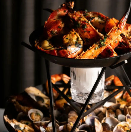 Two-tier seafood tower on a black metal stand with grilled lobster claws and tails glazed in garlic-herb butter above and mixed clams and shellfish on the lower tier — upscale restaurant seafood display.