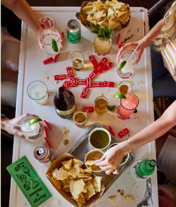 Top-down view of a lively tabletop spread: trays of tortilla chips, bowls of green and red salsa, colorful cocktails and beers, and red dominoes being played as friends reach in.