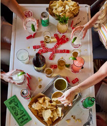 Top-down view of a lively tabletop spread: trays of tortilla chips, bowls of green and red salsa, colorful cocktails and beers, and red dominoes being played as friends reach in.