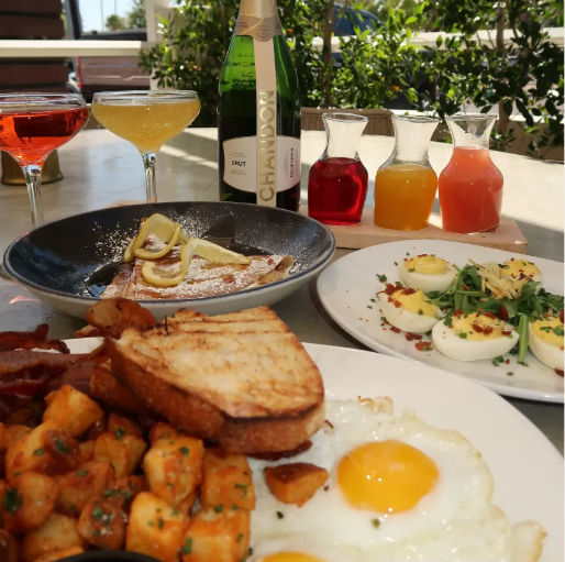 Sunlit patio brunch spread with sunny-side-up eggs, seasoned breakfast potatoes, grilled toast and bacon, deviled eggs with greens, lemon-dusted crepes, sparkling wine bottle and colorful juice and cocktail carafes.