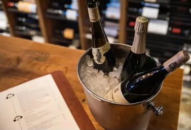Three chilled wine bottles in an ice bucket on a wooden bar counter beside an open wine list, with blurred wine racks in the background — wine bar wine-tasting scene.