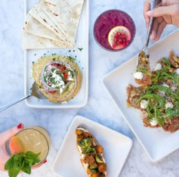 Top-down brunch shot of Mediterranean-style mezze on a marble table: pita with hummus and feta, roasted-vegetable crostini, arugula-topped flatbread, and two colorful cocktails with hands reaching in.
