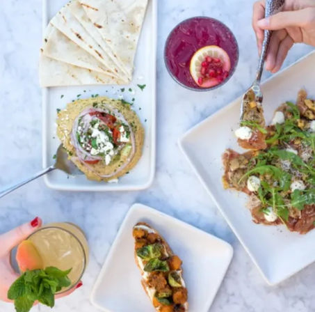Top-down brunch shot of Mediterranean-style mezze on a marble table: pita with hummus and feta, roasted-vegetable crostini, arugula-topped flatbread, and two colorful cocktails with hands reaching in.