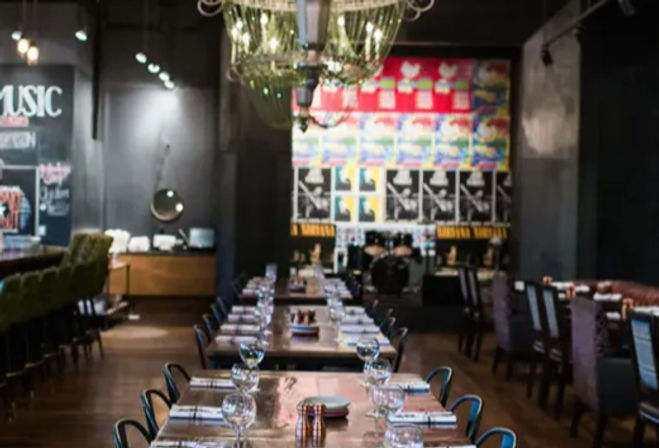 Downtown restaurant interior with a long communal wooden table set with glassware, chandelier overhead, and a small live-music stage with drum kit and colorful posters.