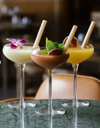 Three colorful cocktails in coupe glasses on a marble table at a cocktail bar — creamy white with an orchid, chocolate-brown with mint and dried lime, and bright yellow with dried citrus, each served with paper straws.
