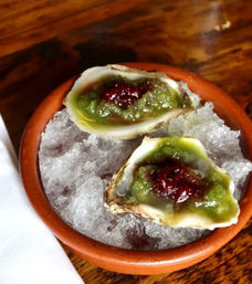 Two oysters on the half shell on crushed ice in a terracotta bowl, topped with bright green sauce and a dollop of red relish — fresh seafood appetizer on a wooden table.
