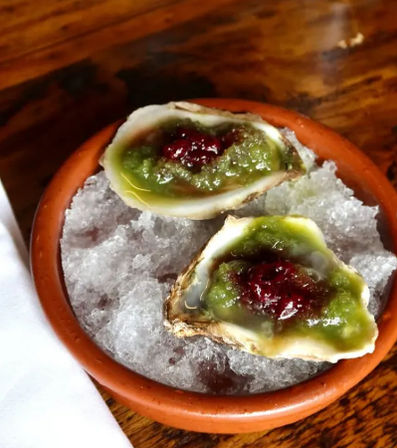 Two oysters on the half shell on crushed ice in a terracotta bowl, topped with bright green sauce and a dollop of red relish — fresh seafood appetizer on a wooden table.