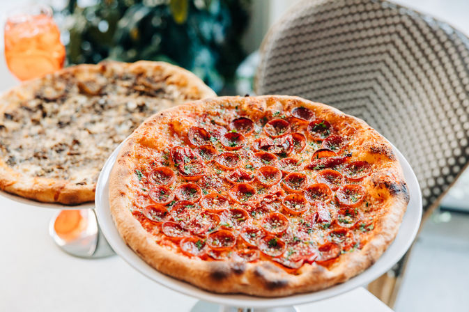 Close-up of a thin-crust pepperoni pizza with curled pepperoni and grated cheese on a white serving stand, a mushroom pizza blurred behind it on a bright pizzeria table.