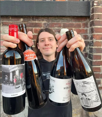 Smiling person holding four assorted craft beer and wine bottles toward the camera with visible labels, standing outdoors against a red brick wall — urban craft beverage showcase.