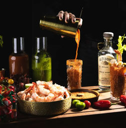 Hand pouring a spicy tomato michelada into a chili-rimmed glass on a dark wooden bar, surrounded by a bowl of shrimp, limes, hot peppers, and green sauces — Mexican-style seafood cocktail setup.