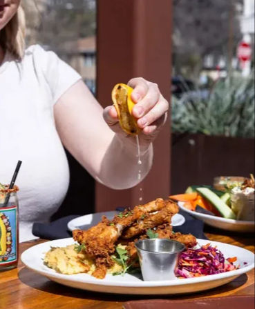 Sunlit outdoor patio: hand squeezing lemon over crispy fried chicken drumsticks on a plate with mashed potatoes, red cabbage slaw and a metal dipping cup.